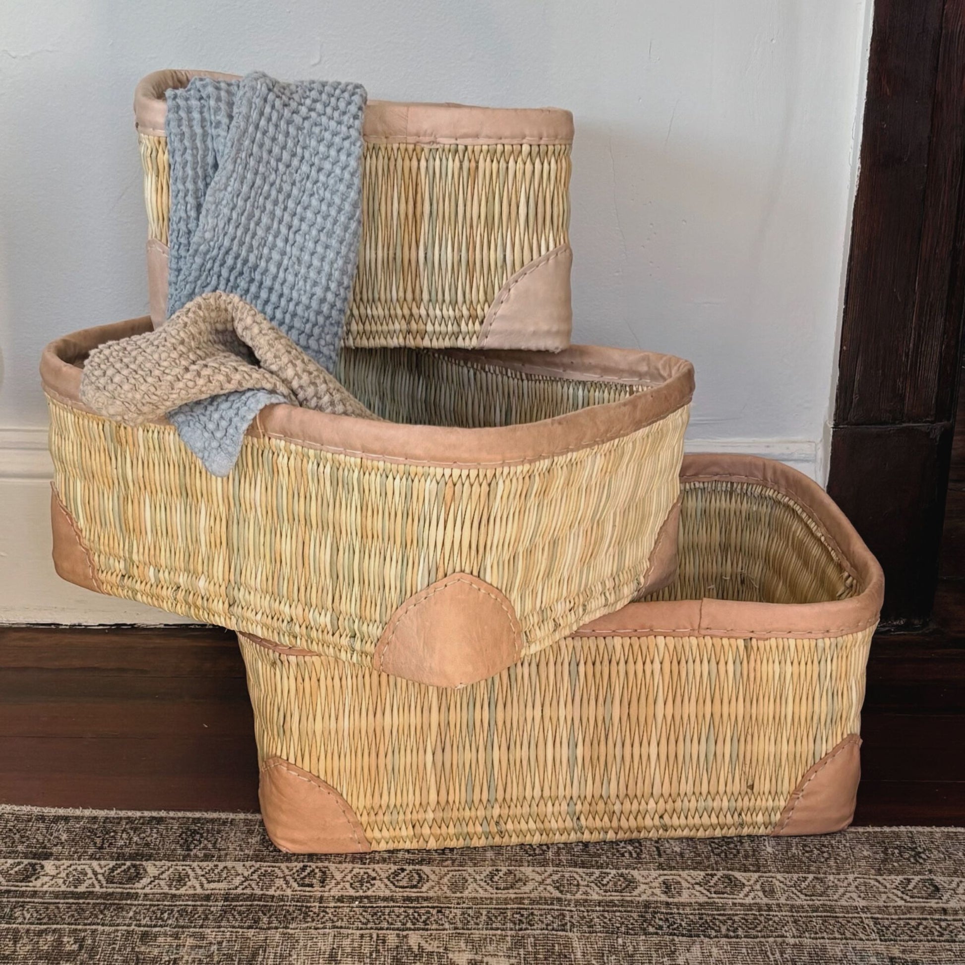 Three stacked rectangular leather-trimmed woven storage baskets on the floor with towels draped over them.