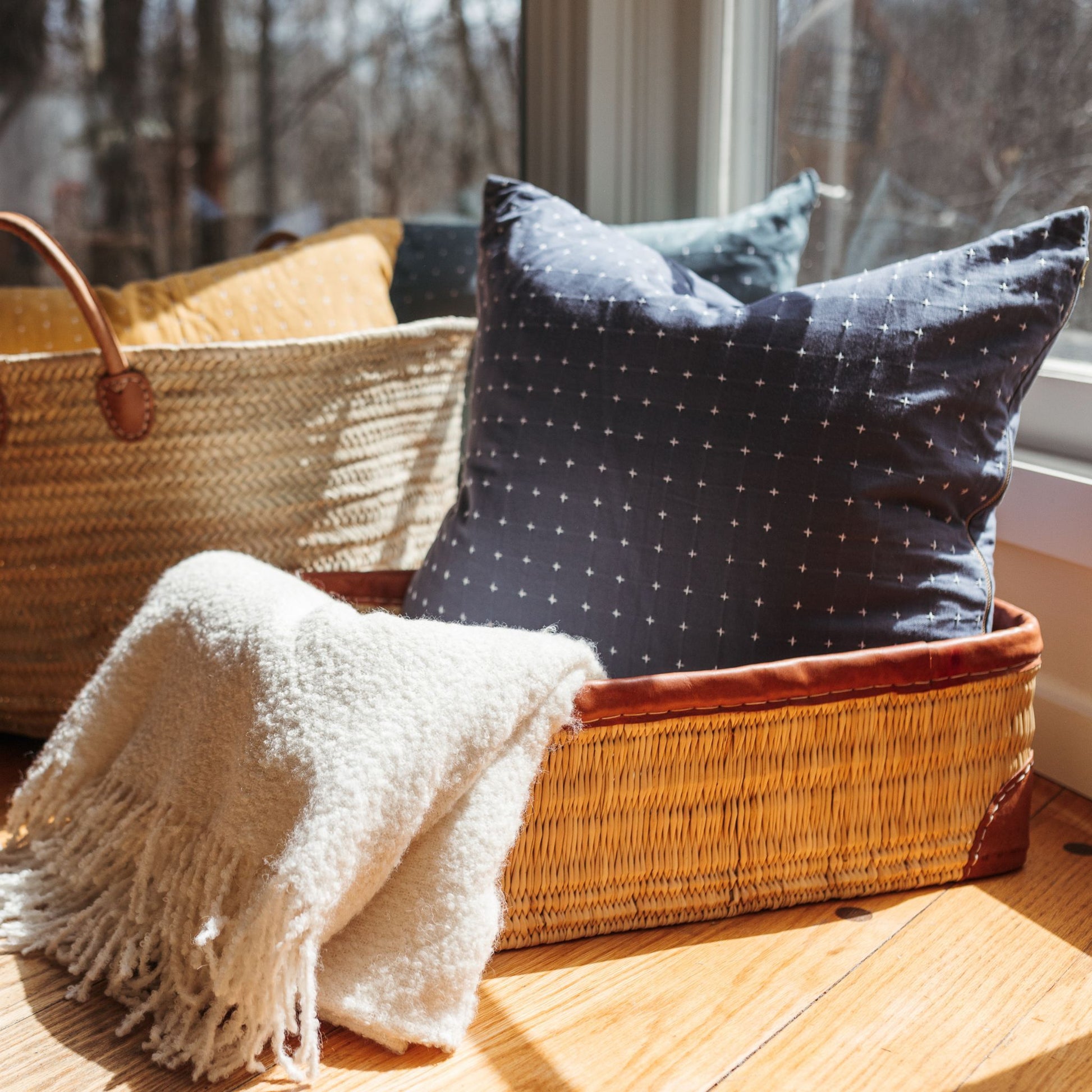 A rectangular leather-trimmed woven storage basket being used to hold a pillow and blanket alongside another basket tote also being used for storage.