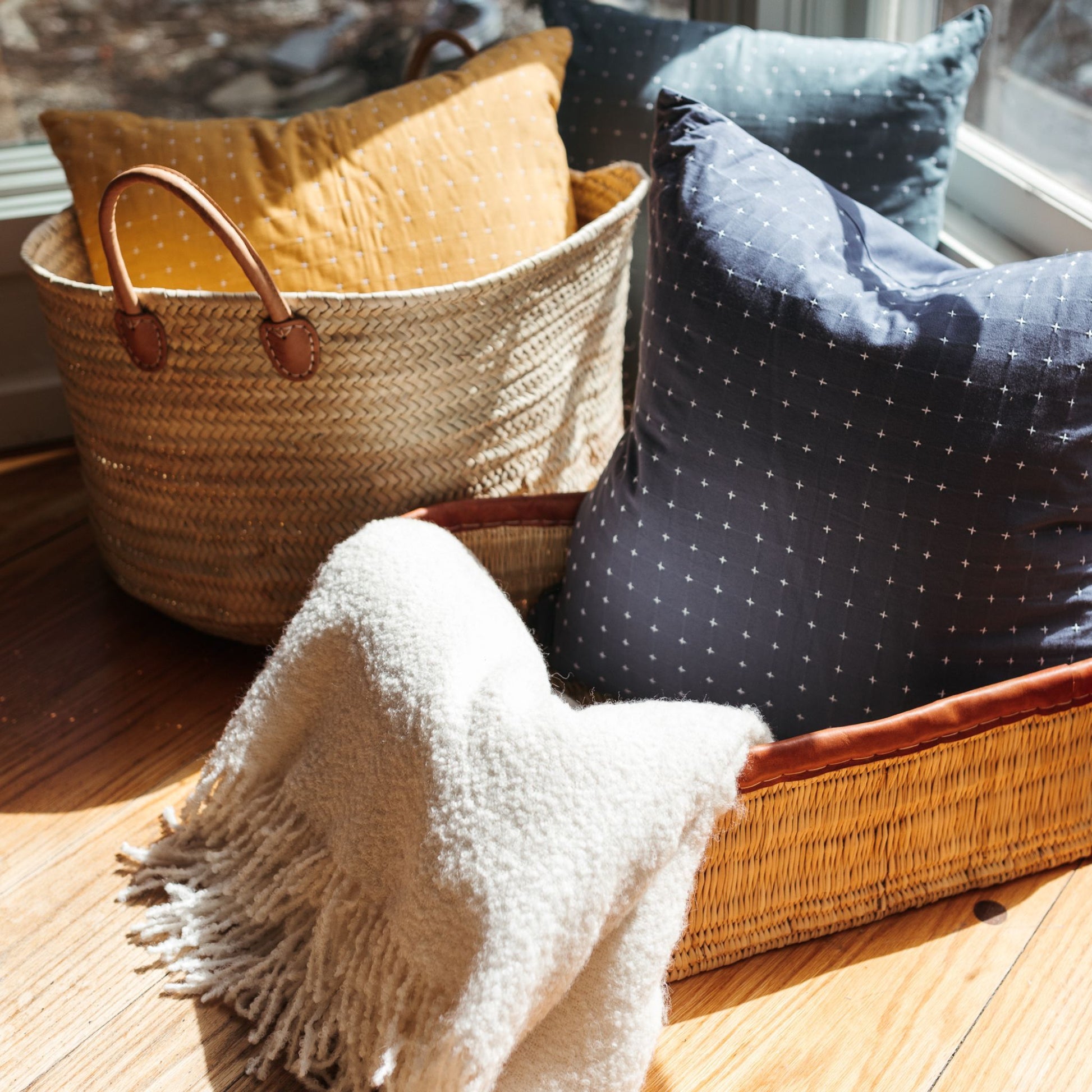 A rectangular leather-trimmed woven storage basket being used to hold a pillow and blanket alongside another basket tote also being used for storage.