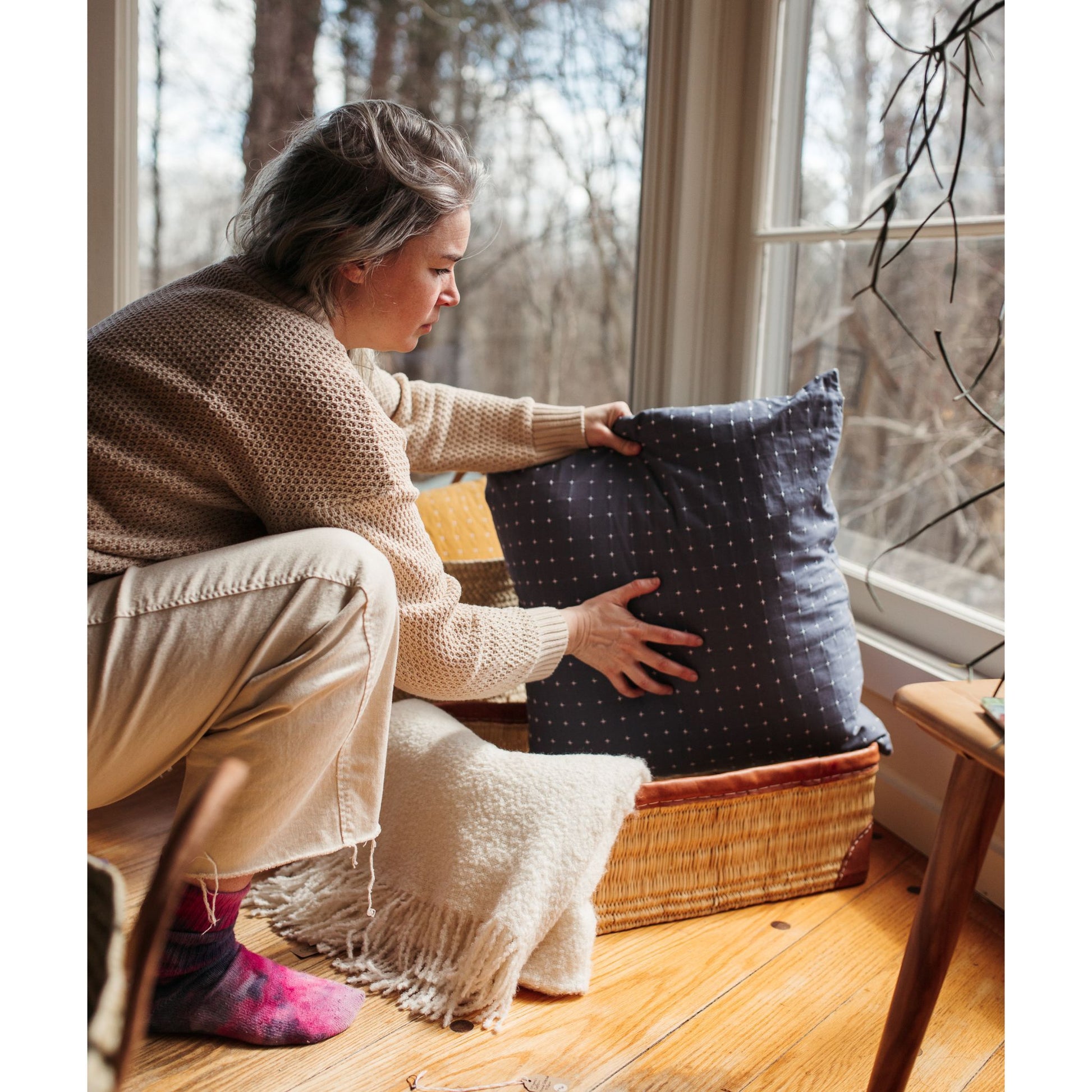 A woman putting a pillow into a rectangular leather-trimmed woven storage basket in a living room.