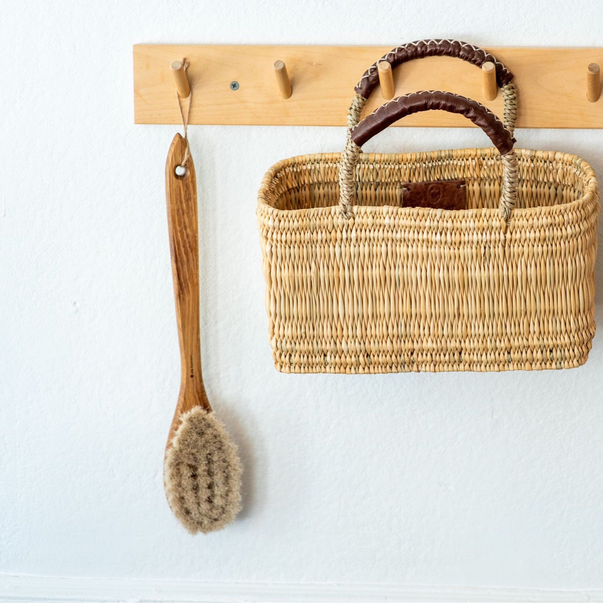 An oak bathing brush with natural fiber bristles hanging from a wall rack alongside a rectangle basket