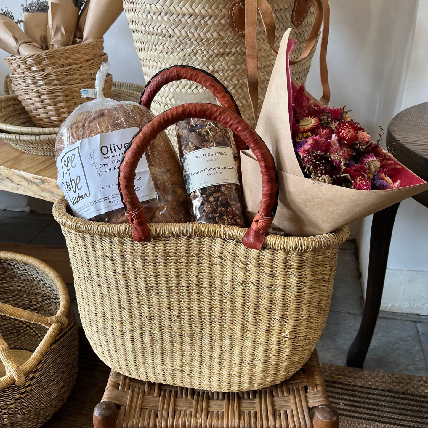 A woven, oval bolga tote basket with leather handles holding a bread loaf, granola, and a dried flower bouquet.