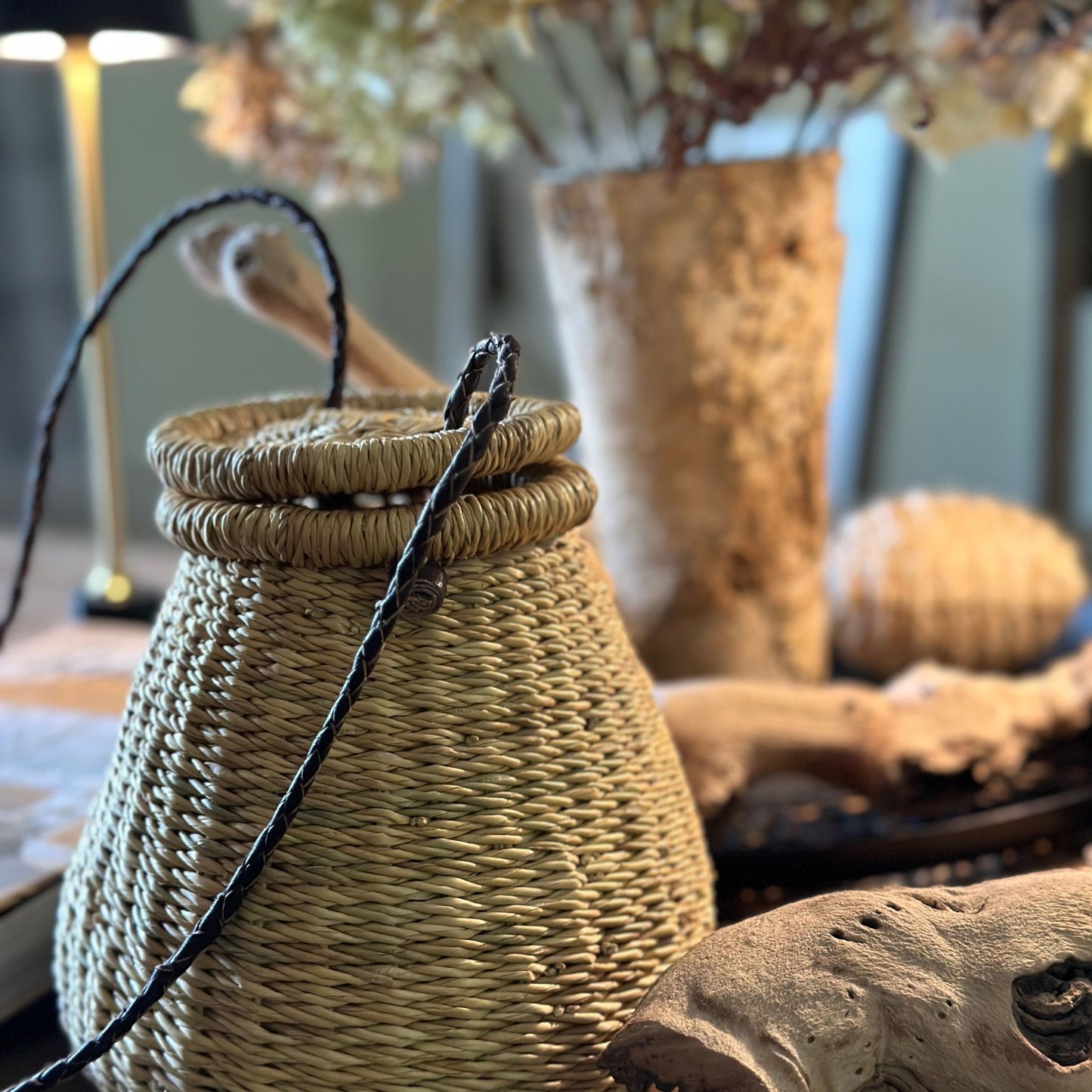 Lidded crossbody pear-shaped basket bag with a brown leather braided strap and a lamp and vase with hydrangeas in the background.