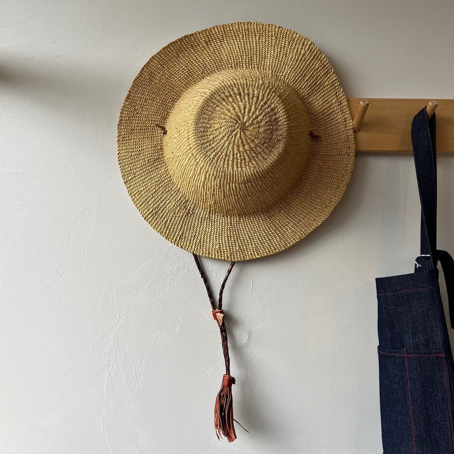 A tan, short brim straw sun hat with a braided strap hanging on a wall hook next to a denim apron.