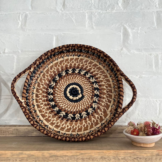 Overhead view of shallow, round, pine needle tray basket with handles. It has a brown, black, and tan woven design on the top. The background is white brick and the basket is on a wooden shelf with dried flowers alongside it.