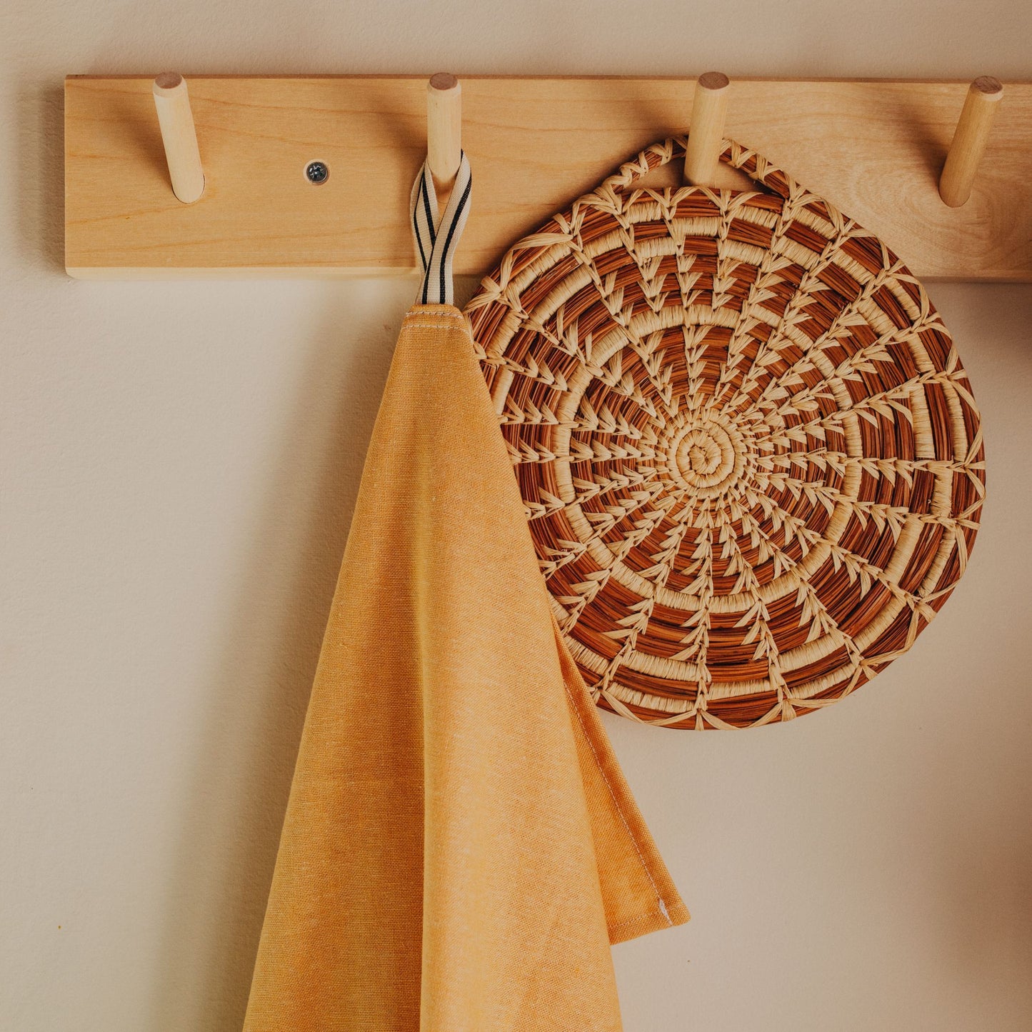 An 8" brown and tan pine needle woven trivet hanging on a wall hook next to a yellow hand towel.