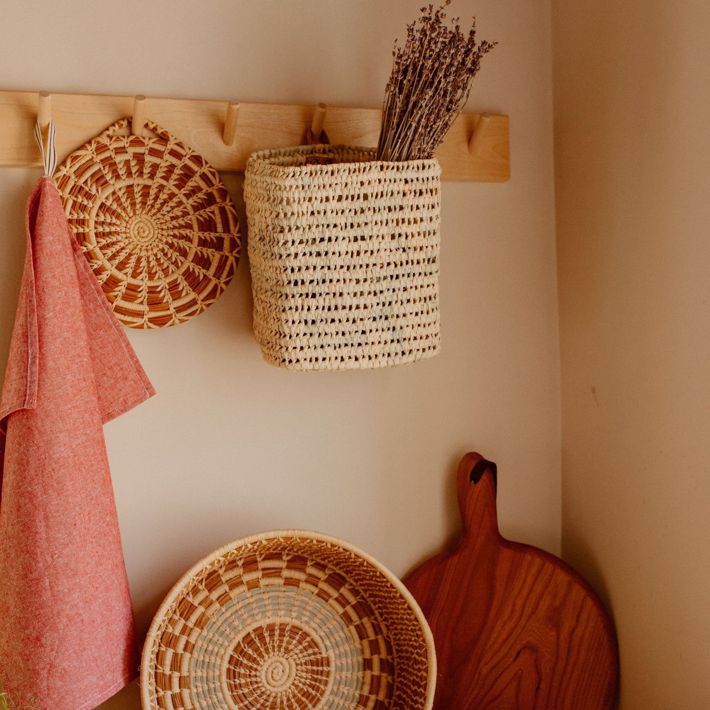 A square hanging basket hanging from a wall hook with dried lavender inside of it. Next to the basket is a trivet and towel. On the counter below them is another round basket and wooden cutting board.