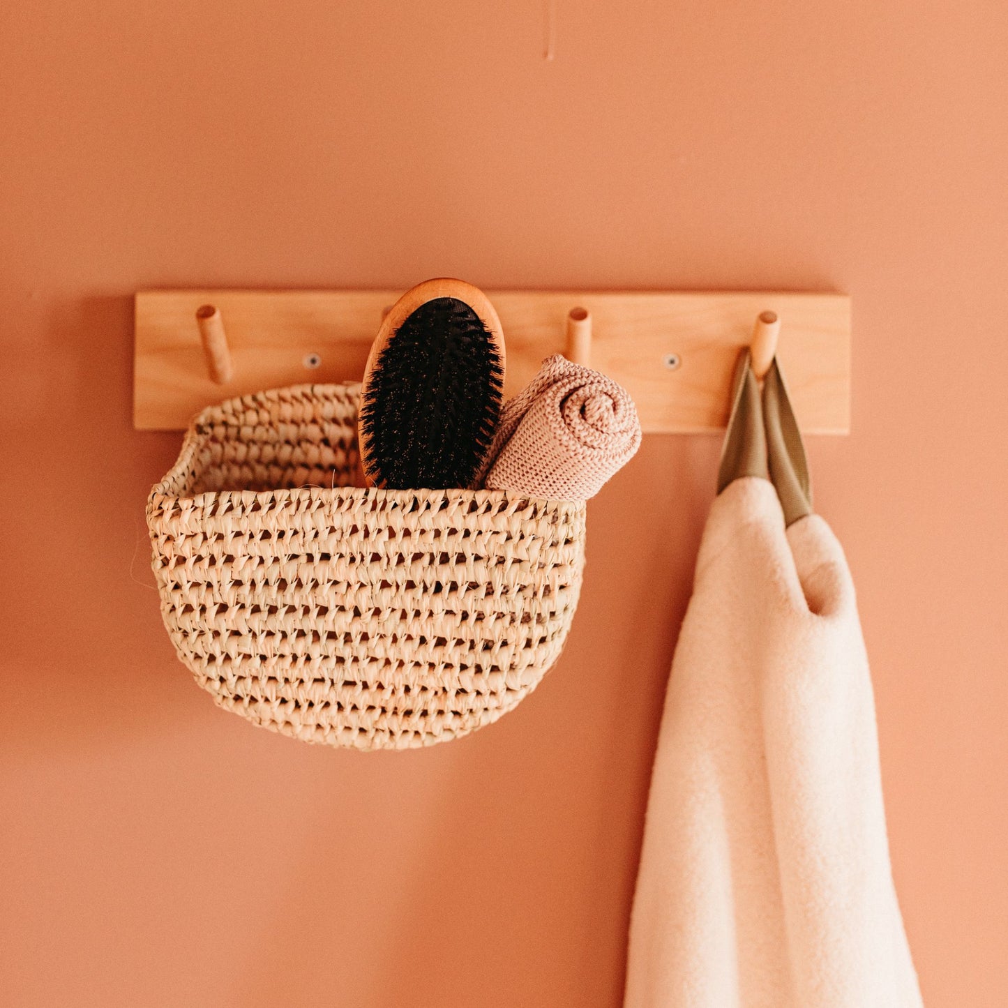 A rounded hanging basket on a wall hook holding a hair brush and face towel alongside another hanging towel.