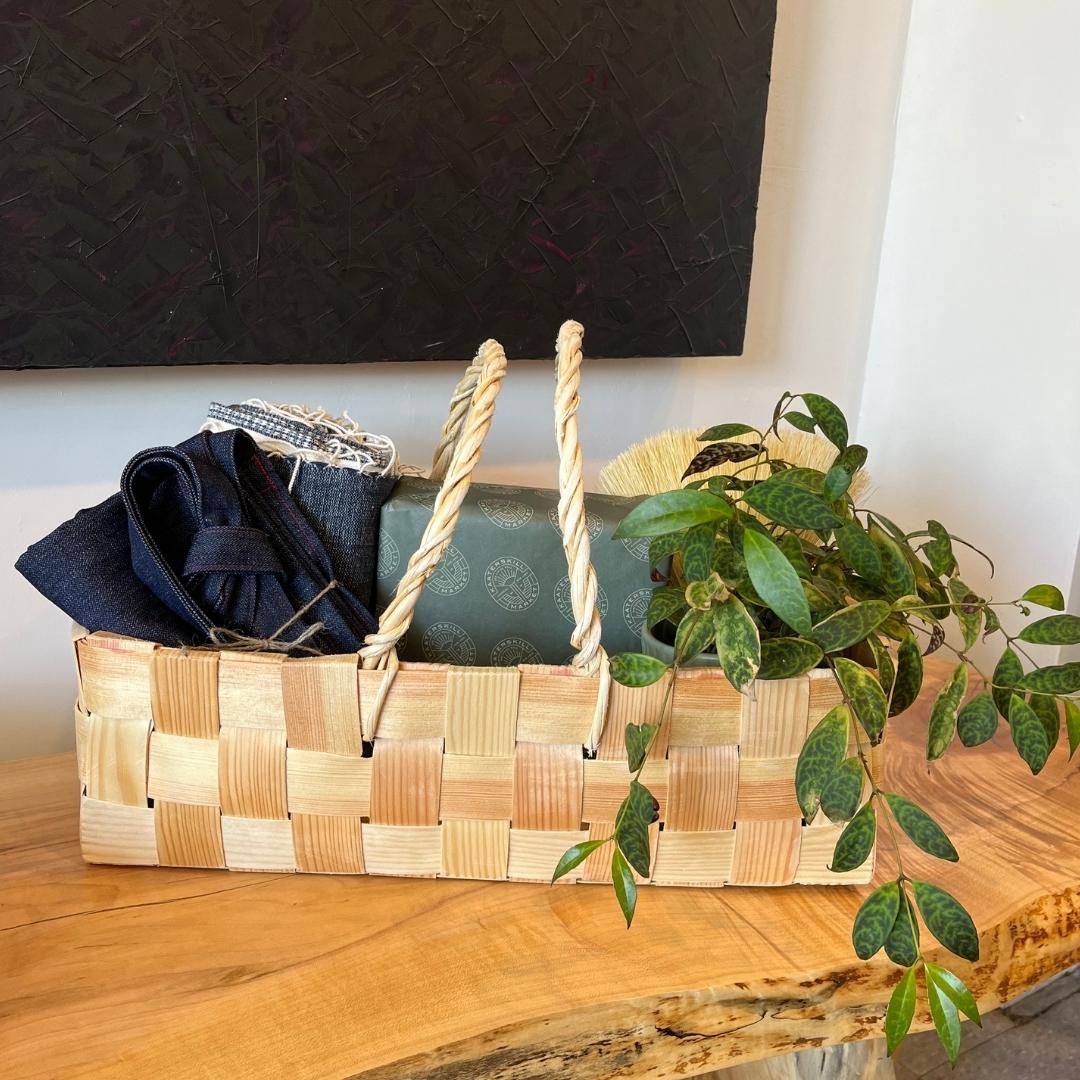 A traditional pine chipboard basket with handles holding napkins, an apron, a wrapped gift, and a plant on a wooden bench.