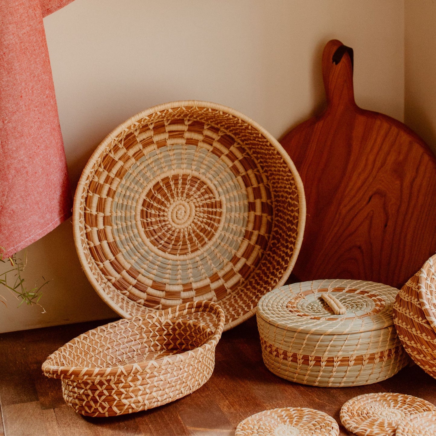 A kitchen counter with three pine needle baskets. The harvest basket is in the center besides a cutting board and a red hand towel hanging from the wall and pine needle coasters are in front of the baskets.