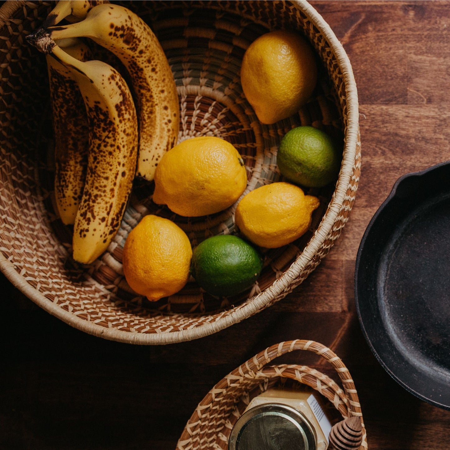 A brown, tan, and light green handwoven pine needle harvest basket holding limes, lemons, and bananas next to a skillet and a smaller pine needle basket holding honey.