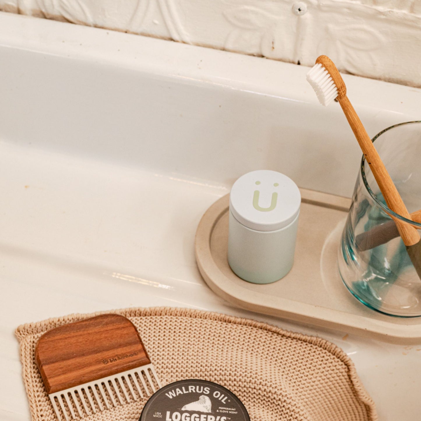 A toothpaste tin next to a glass cup with two toothbrushes on a tray on a bathroom counter. Next to the tray is a washcloth with a comb and a "Walrus Oil" product.