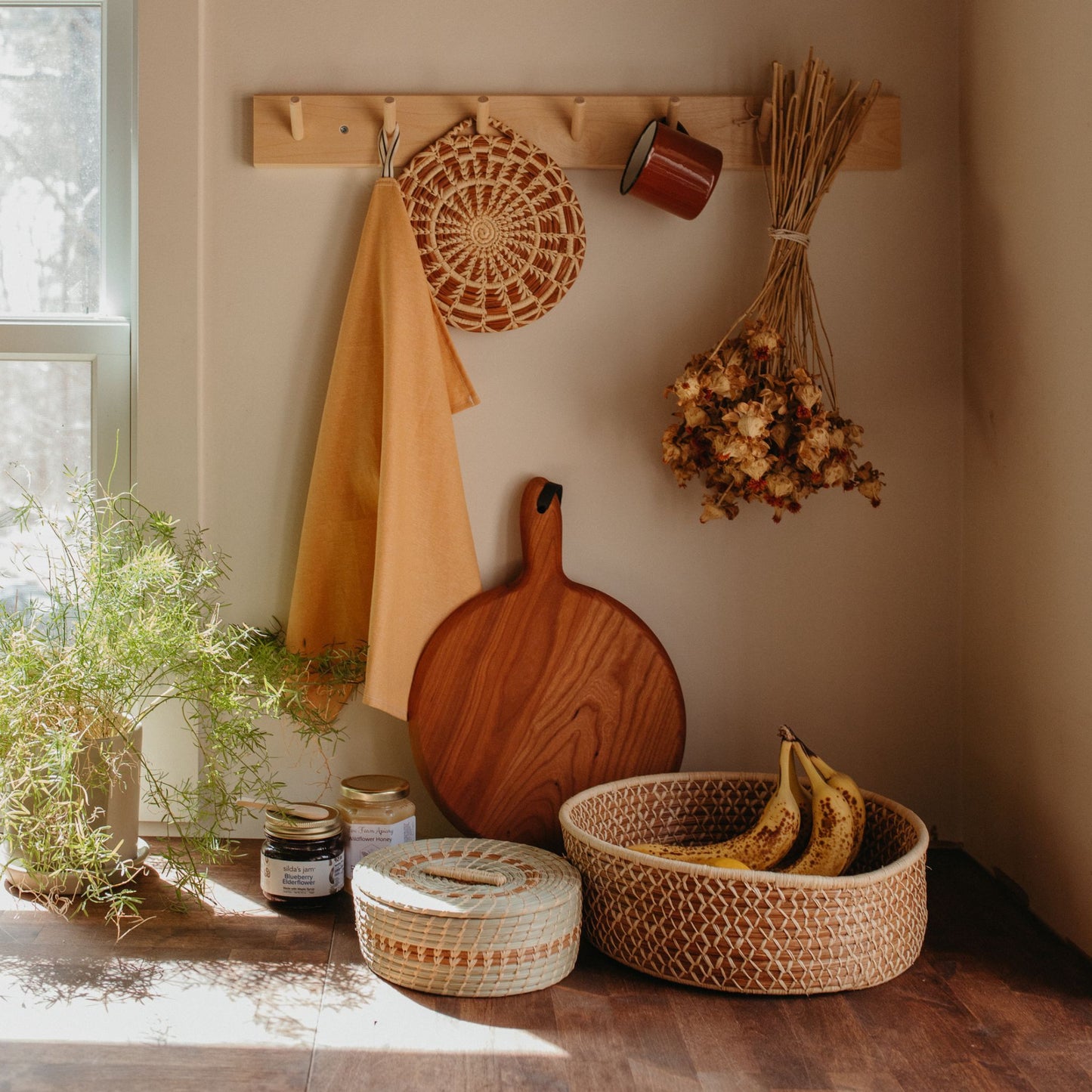 A pine needle woven trivet hanging on a wall hook next to a yellow hand towel, mug, and bouquet of dried flowers. Leaning against the wall is a cutting board and a plant, the tortilla basket is in the middle next to a larger basket filled with fruit.