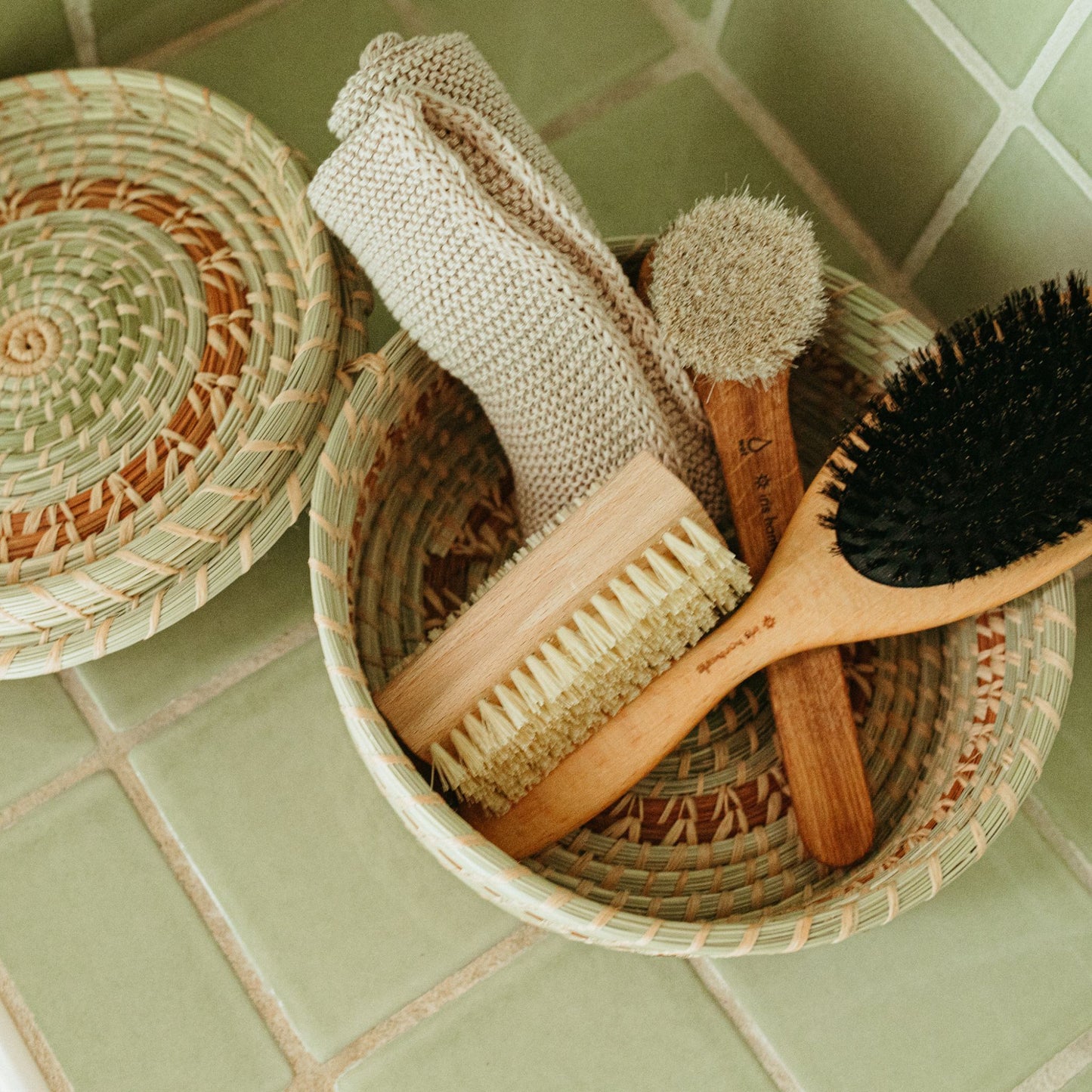 A green and brown 8" x 2.5" lidded basket filled with a towel, face brush, hair brush, and nail brush with a green tile background.