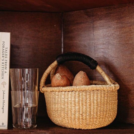A mini round bolga basket with a black leather handle holding vegetables next to glassware.