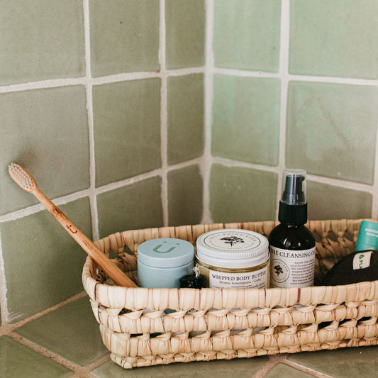A basket filled with a toothbrush, a refill tin, body butter, a cleanser, and face washing rounds in front of green tile.