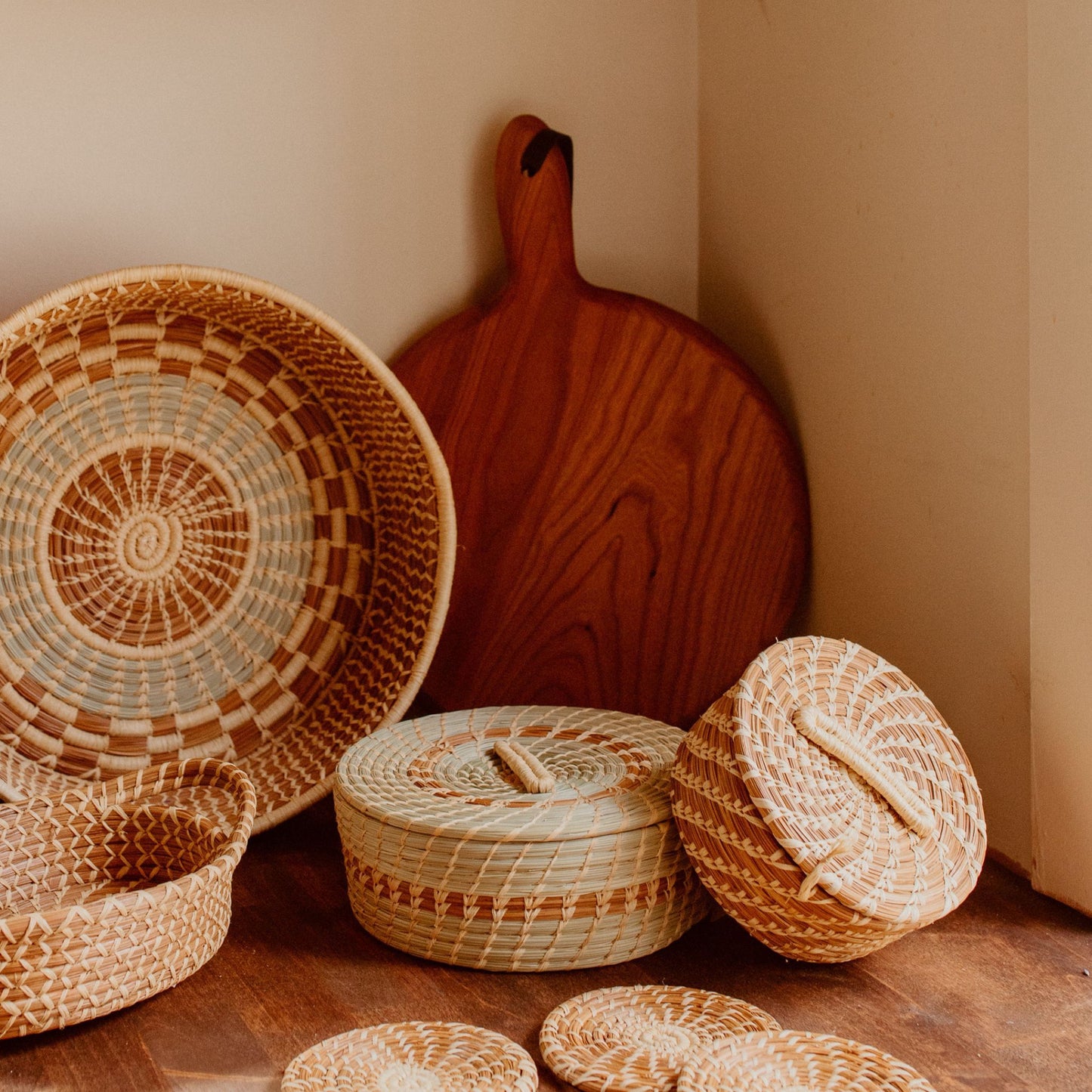 Small woven pine needle lidded basket leaning against green tortilla basket among larger baskets and a cutting board.