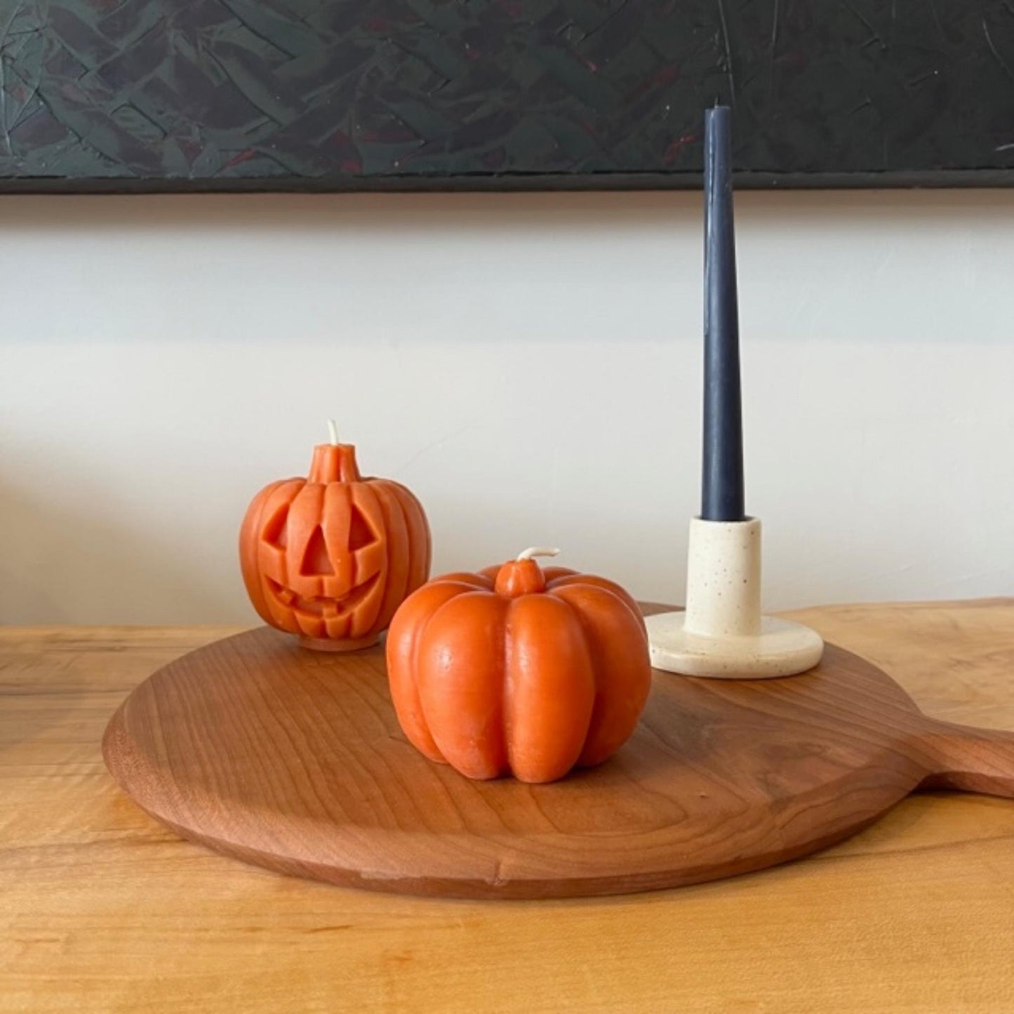 2 beeswax pumpkin candles sit on round wood cutting board next to a taper candle in ceramic candlestick