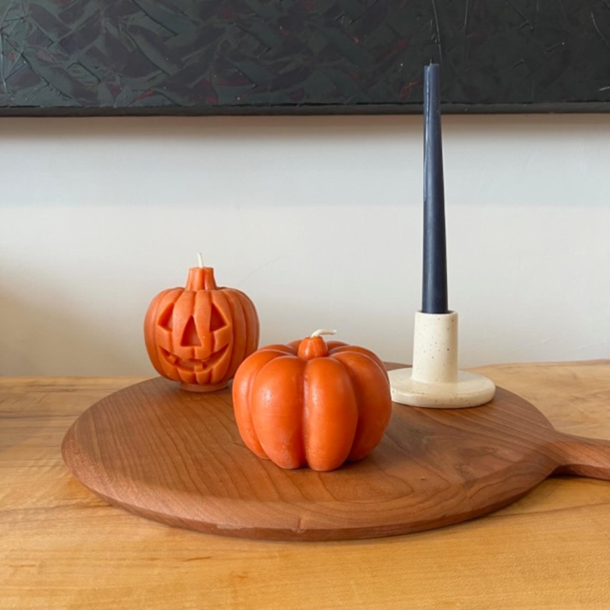 2 beeswax pumpkin candles sit on round wood cutting board next to a taper candle in ceramic candlestick