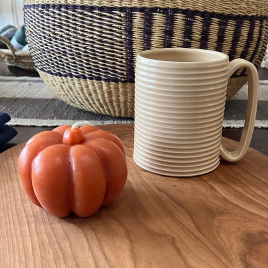 Hand-poured beeswax pumpkin candle sits on a wooden table next to a tall mug