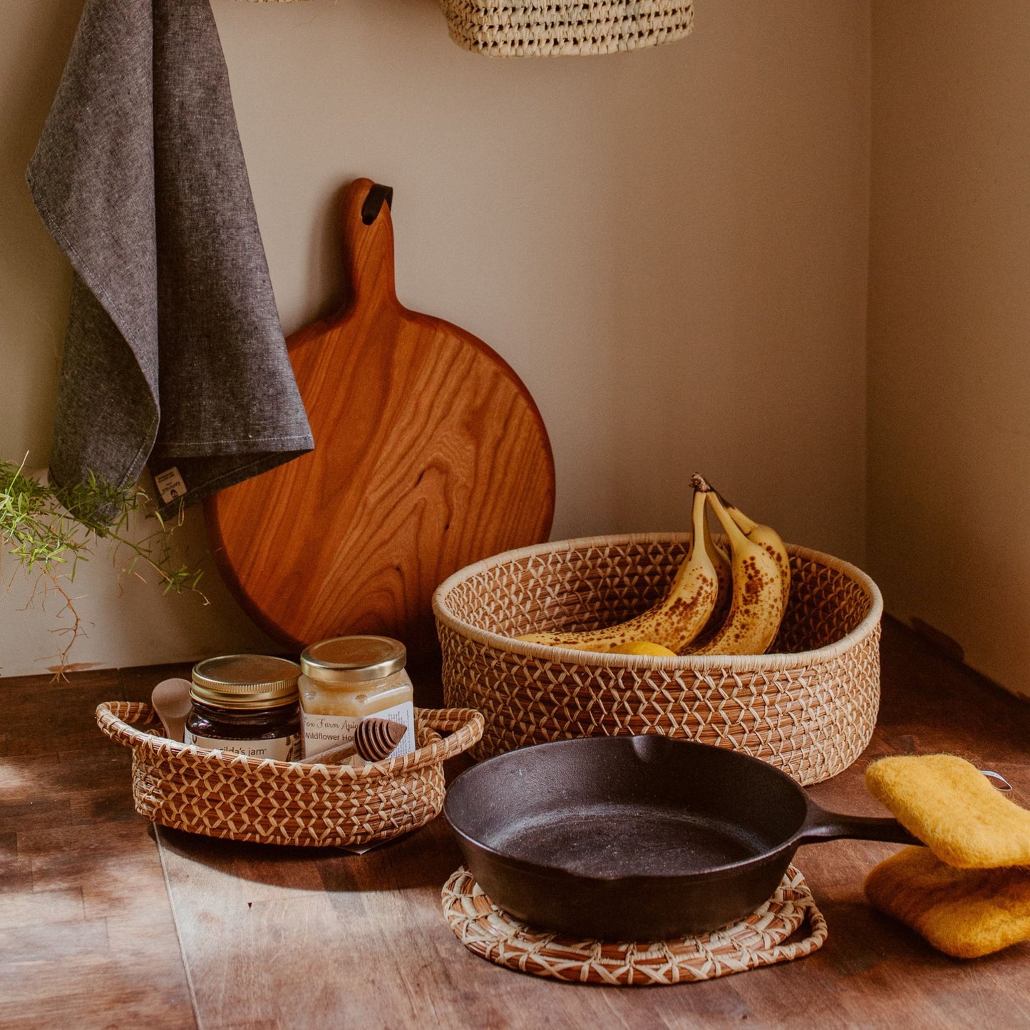 A kitchen counter with a cutting board, two baskets filled with fruit and honey, a towel hanging from the wall and a skillet using the pine needle trivet with a yellow pot holder.