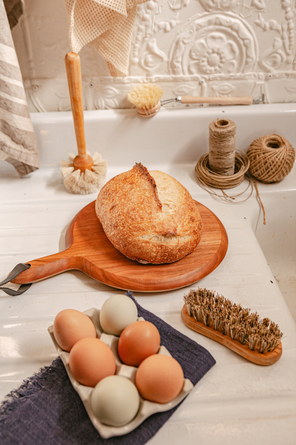 A vegetable scrub brush on a kitchen sink alongside a loaf of sourdough and eggs. In the background are dish brushes, twine, and hanging towels.