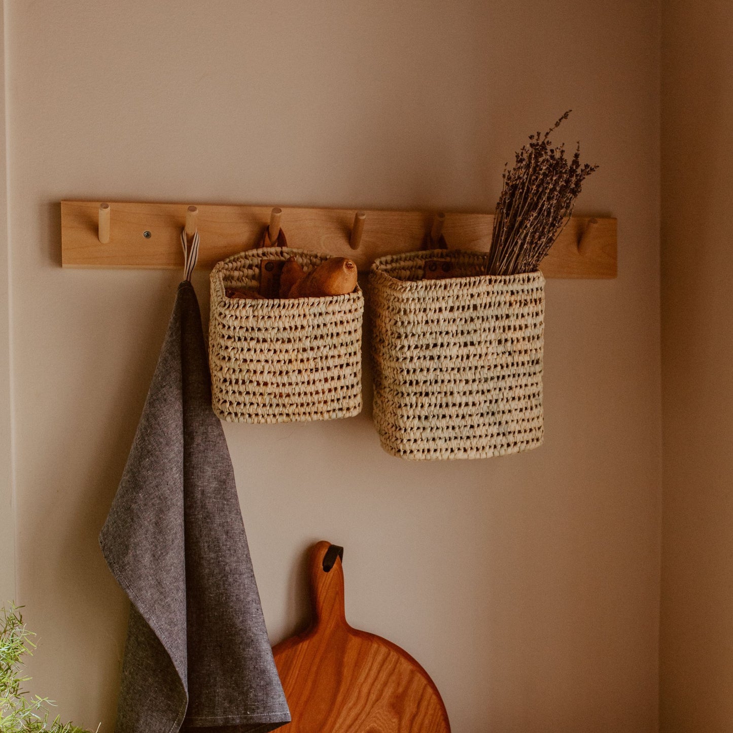 A square hanging basket hanging from a wall hook with dried lavender inside of it. Next to the basket is another square basket and towel. On the counter below them is a wooden cutting board.