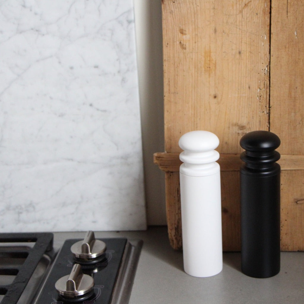 A black and a white salt or pepper mill grinder made of beechwood, positioned on a kitchen countertop.