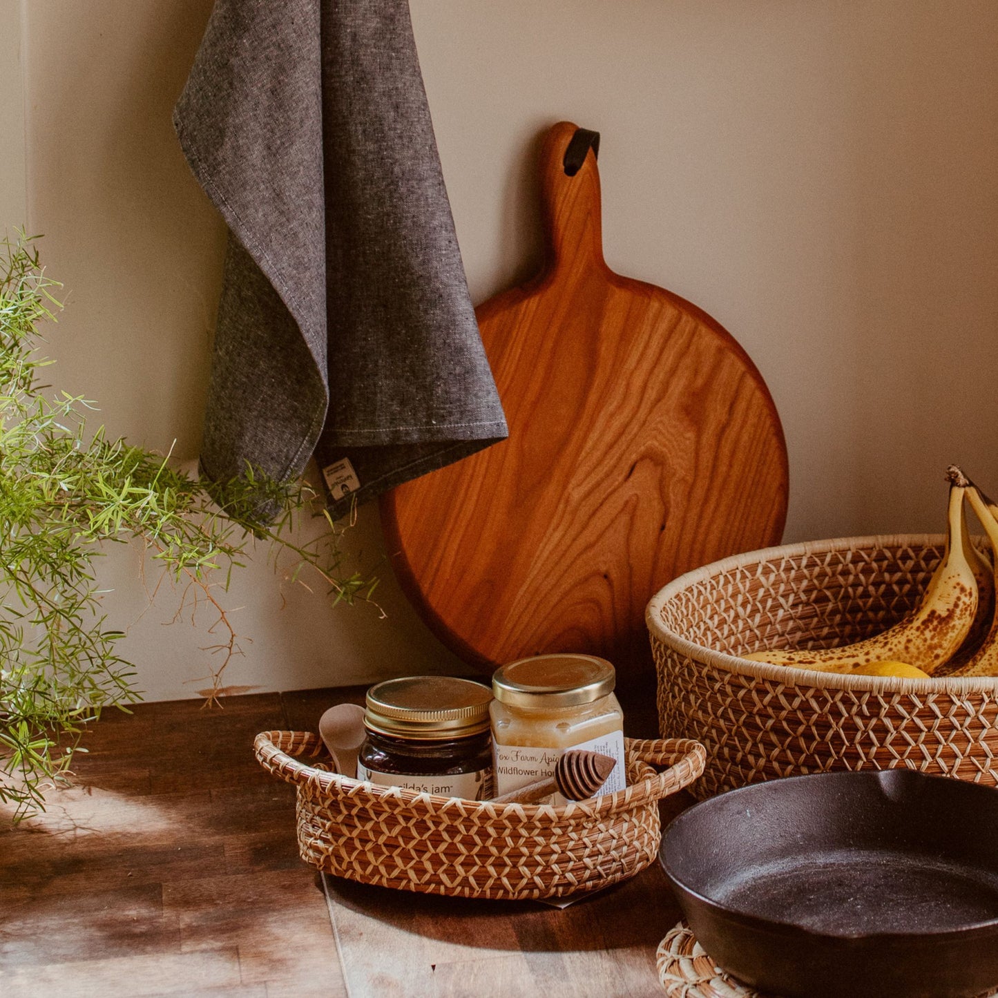 Pine needle basket with handles filled with honey and jam on a kitchen counter. In the background is a cutting board, hanging towel, larger basket with fruit, a plant, and a skillet alongside the basket.