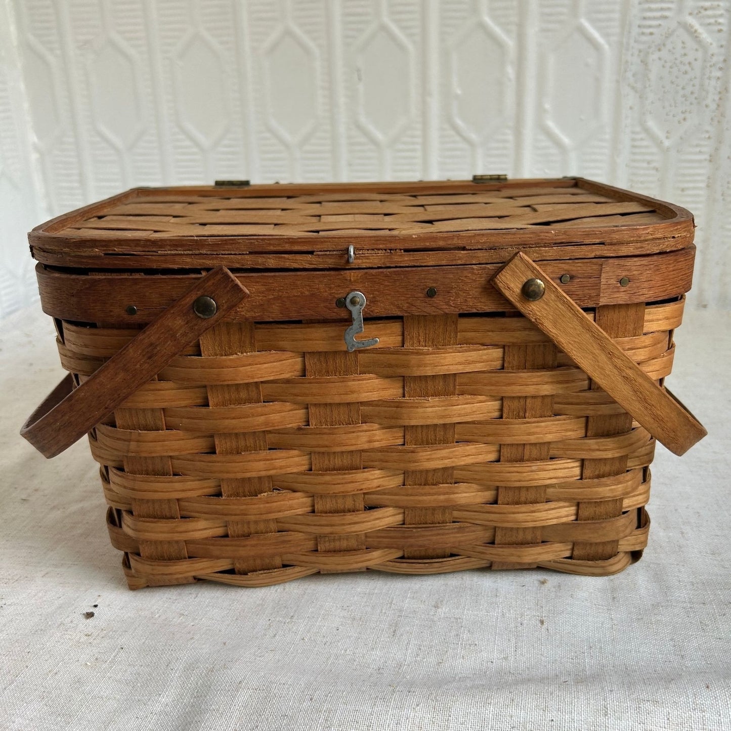 A vintage rectangular wovenpicnic  basket with a slatted wooden lid, two wooden side handles, and a small metal latch on the front, photographed against a white textured backdrop.