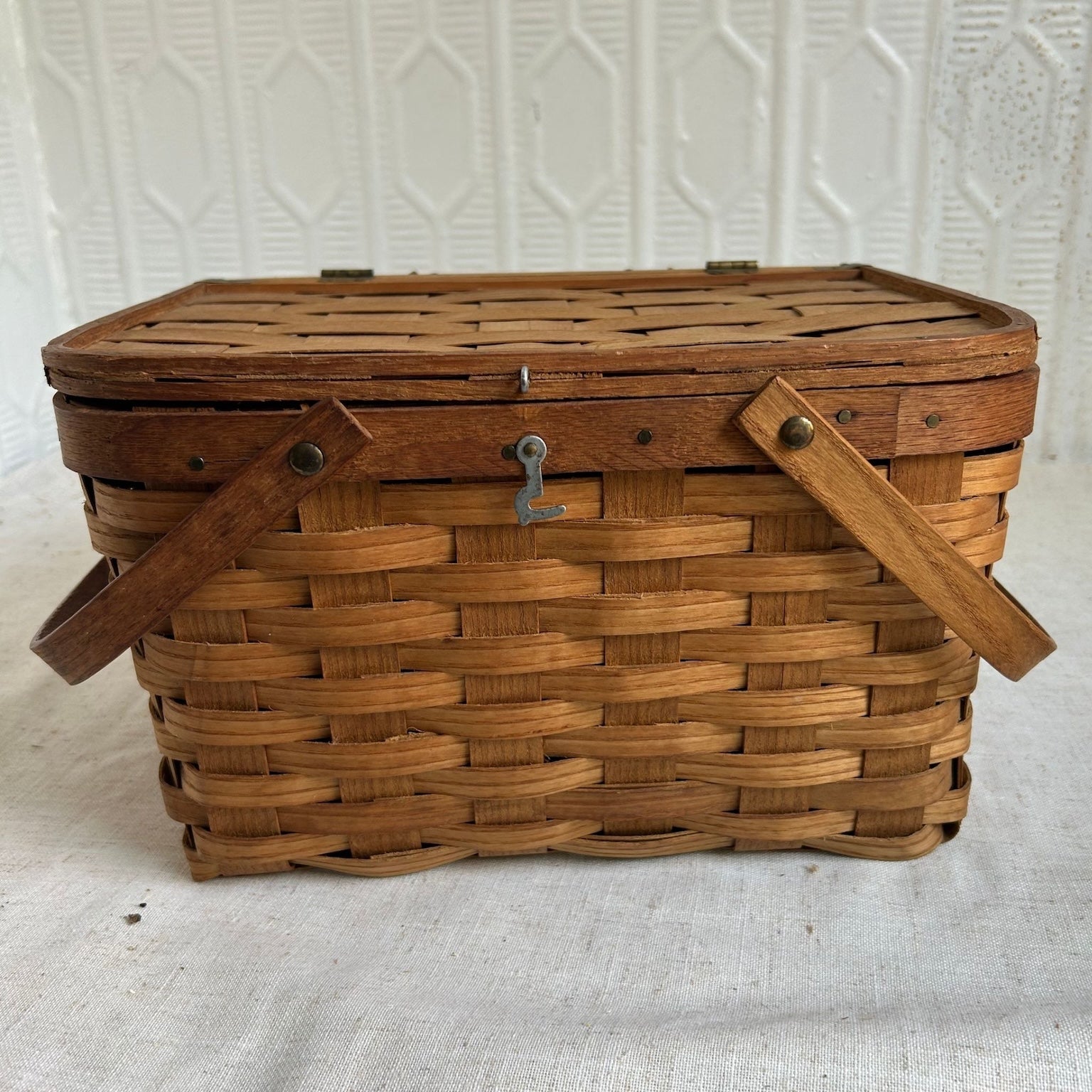 A vintage rectangular wovenpicnic  basket with a slatted wooden lid, two wooden side handles, and a small metal latch on the front, photographed against a white textured backdrop.