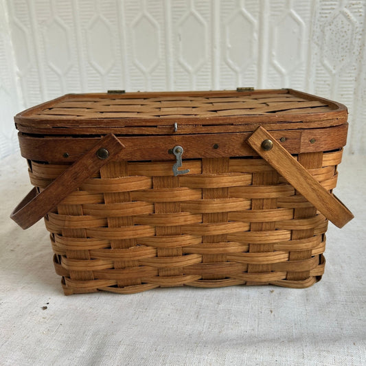 A vintage rectangular wovenpicnic  basket with a slatted wooden lid, two wooden side handles, and a small metal latch on the front, photographed against a white textured backdrop.