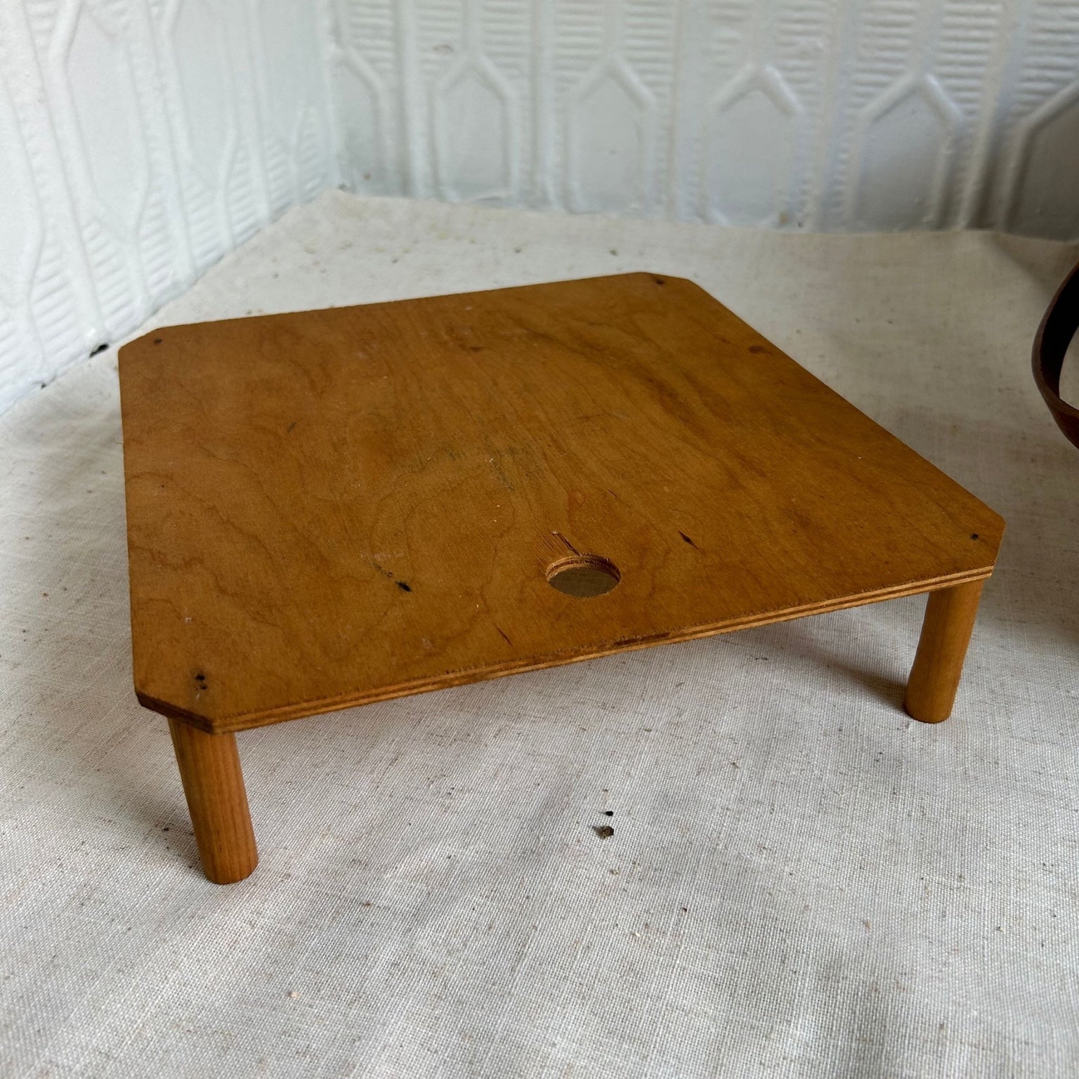 A small square wooden table or stand with four legs and a circular hole in the top surface, photographed on a light fabric surface against a white textured wall.