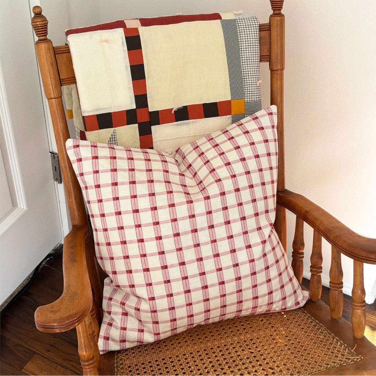 A red and white gingham checkered throw pillow resting on a wooden rocking chair with a patchwork quilt draped over the back.