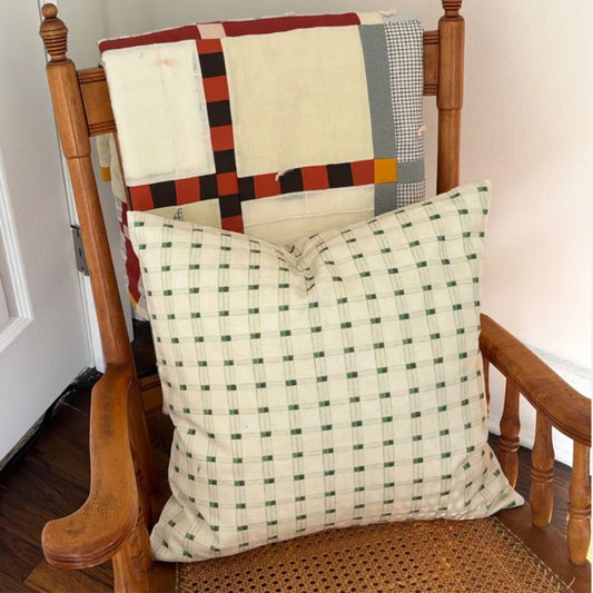 A square throw pillow with a green and cream grid pattern is styled on a wooden rocking chair, with a multicolor patchwork quilt draped over the backrest.