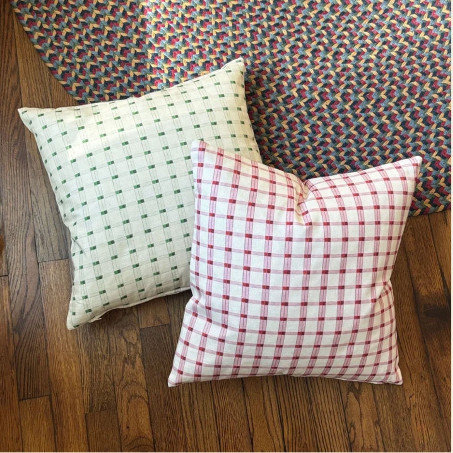 Two square throw pillows with gingham patterns, one in green on an off-white background and the other in red on a white background, displayed on a wooden floor with a multicolored woven rug behind them.