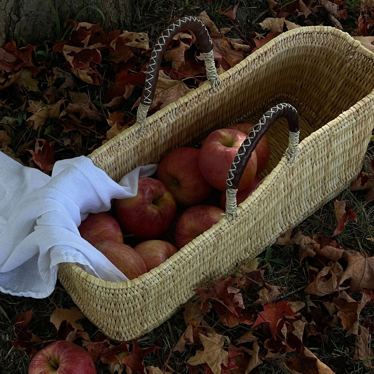 A rectangular woven basket with leather handles holding apples placed on autumn leaves. 