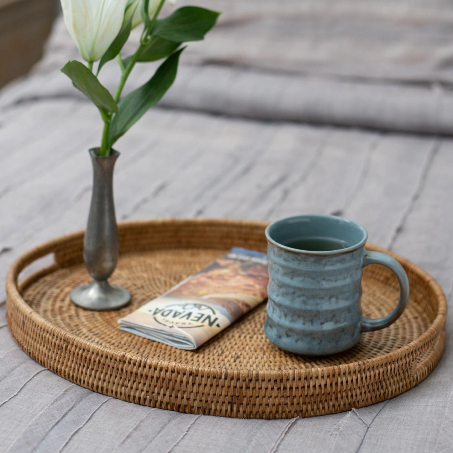 Wicker tray with a blue mug, vase with white tulip, and newspaper on a wooden surface.