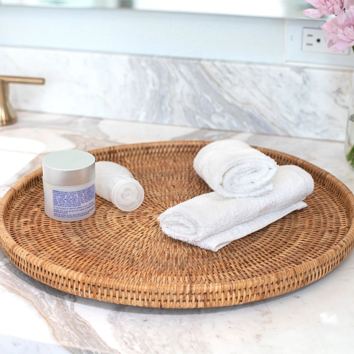 Round woven tray with a jar, rolled towels, and folded towel on a marble countertop.