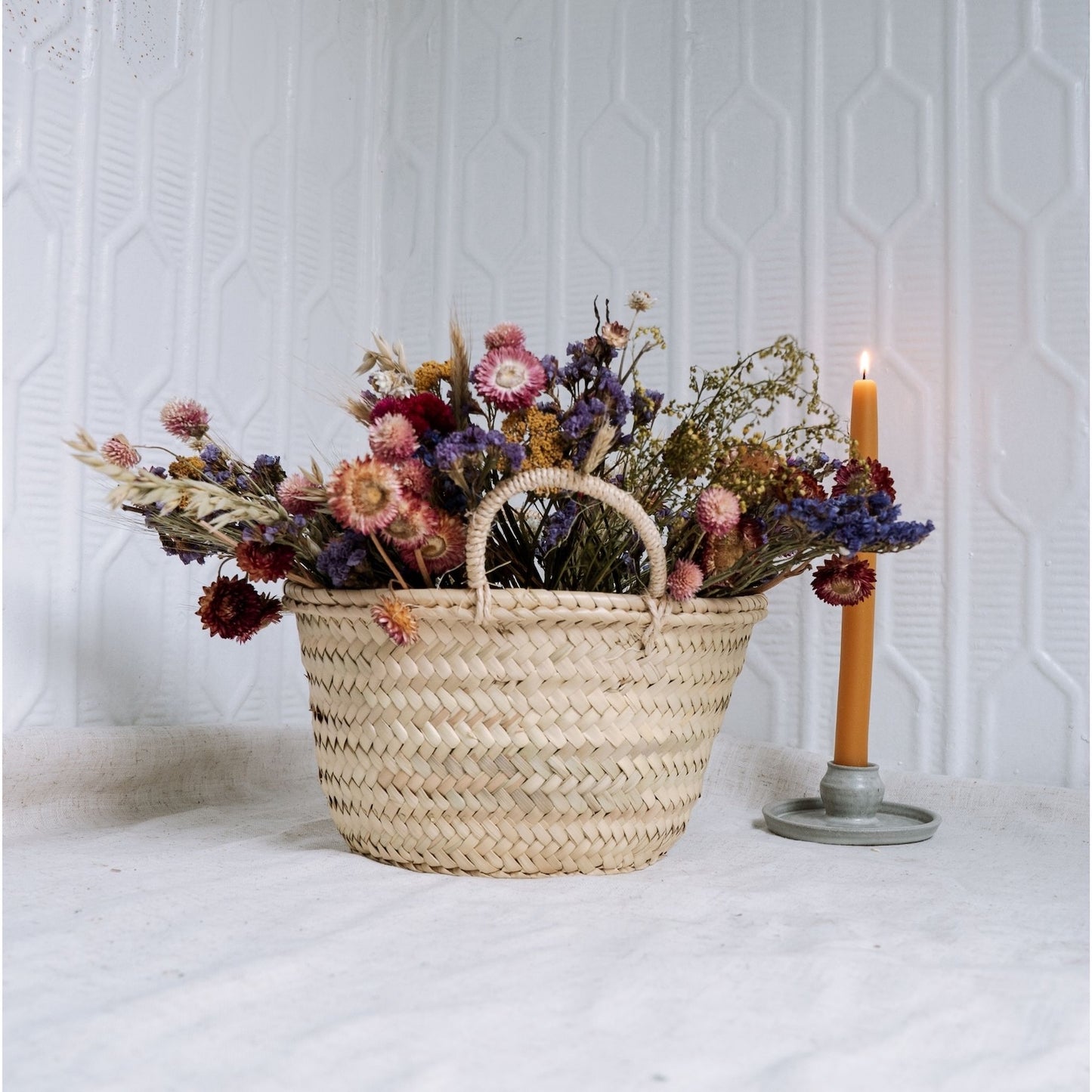 Woven basket with dried flowers and a lit candle on a white surface.