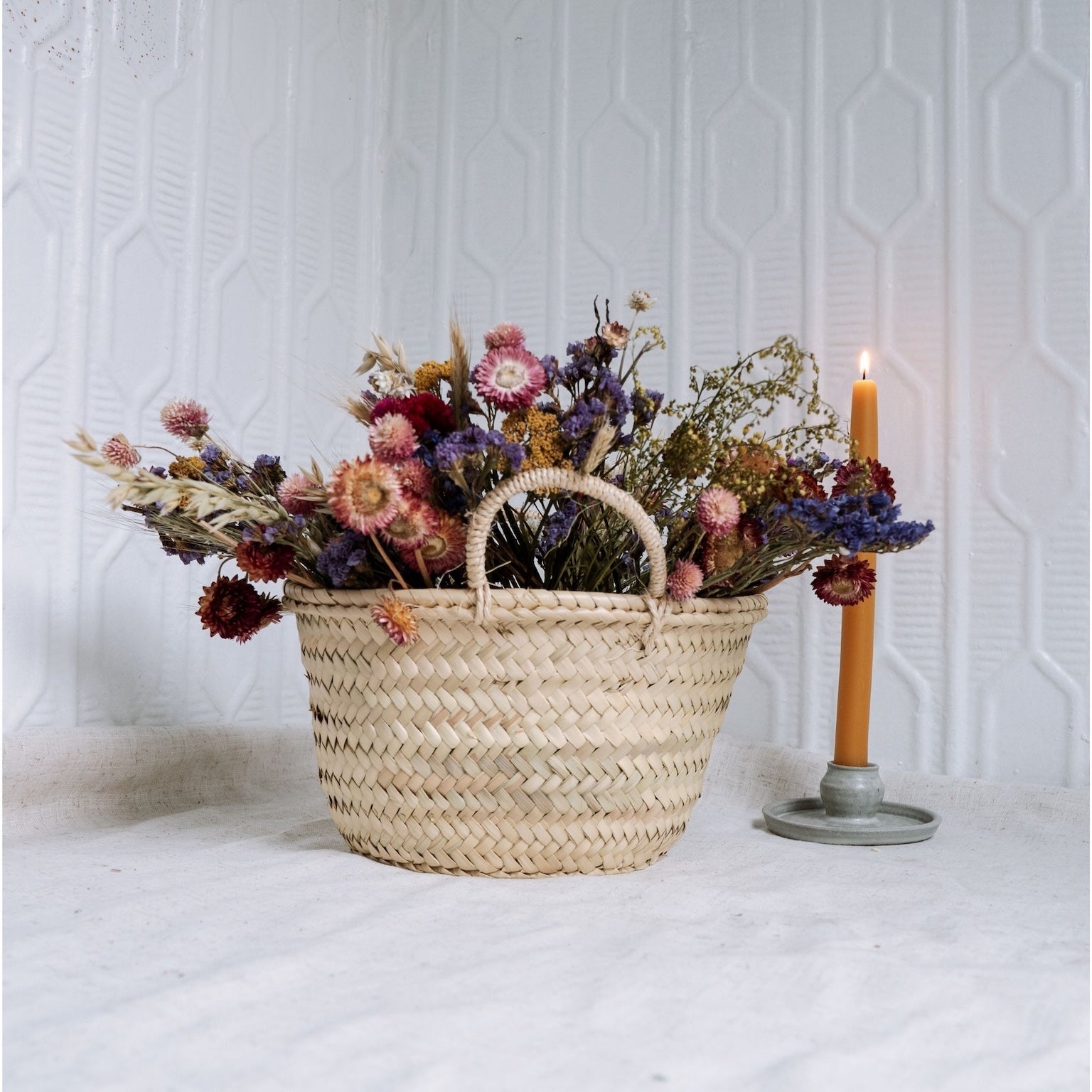 Woven basket with dried flowers and a lit candle on a white surface.