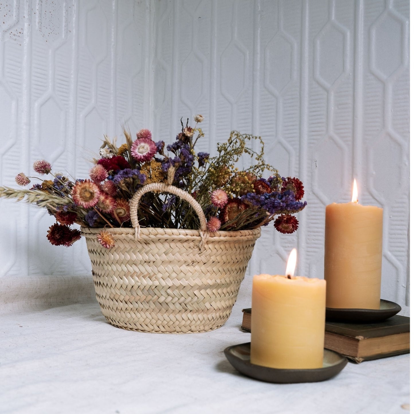 Woven basket with dried flowers and two candles on a textured white surface