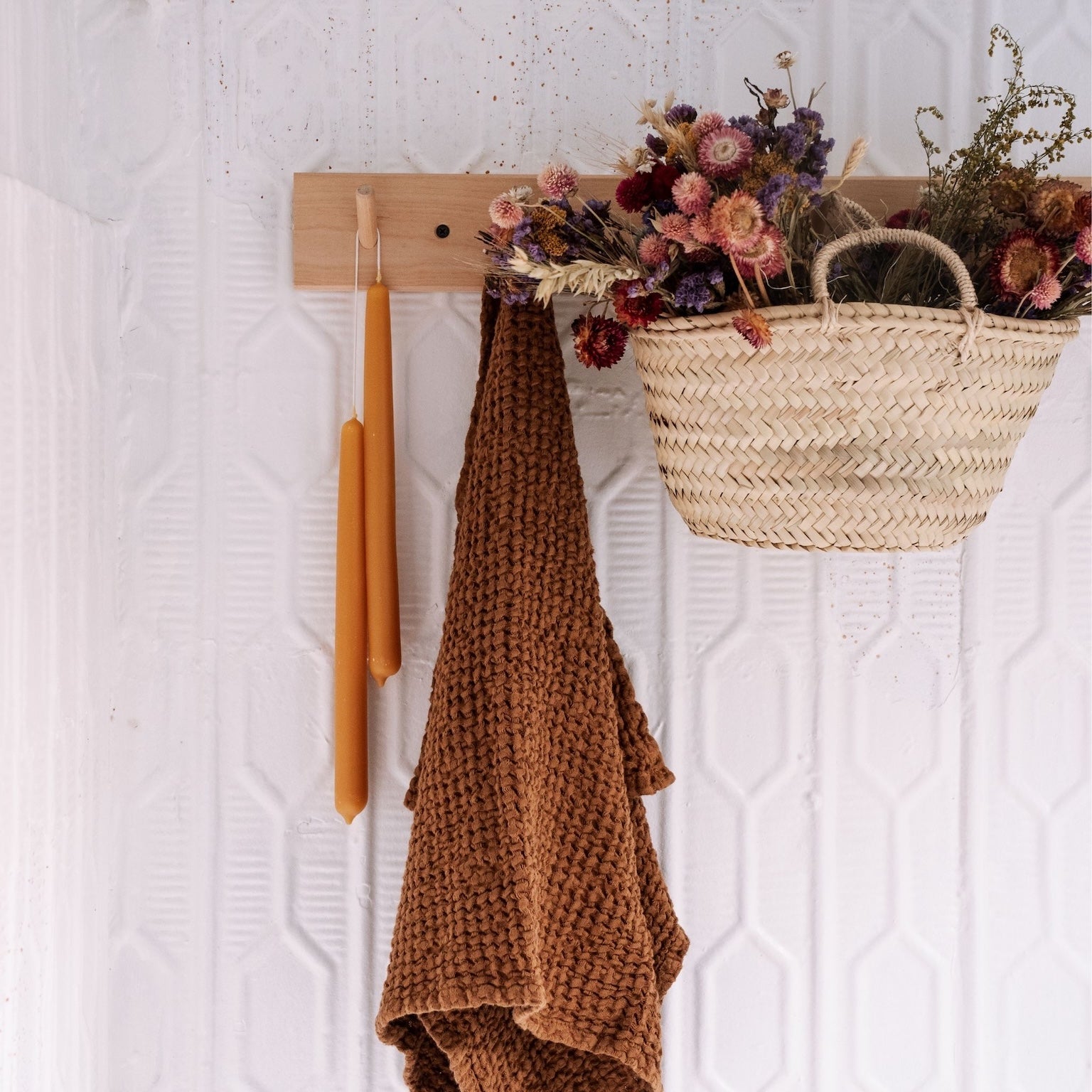 Decorative wall hook with a brown linen towel, two beeswax candles, and a woven basket with flowers on a textured white wall.