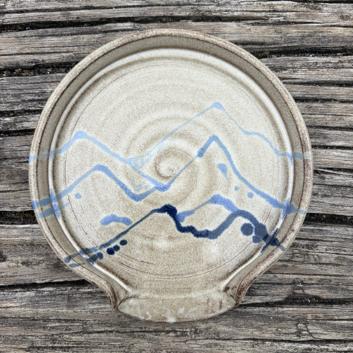 A round handmade ceramic spoon rest with a textured beige glaze, featuring a blue painted mountain and river pattern, displayed on a rustic weathered wood background.