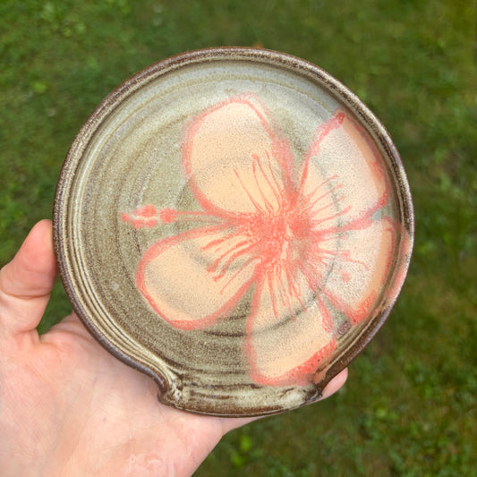 A hand holds a round handmade ceramic spoon rest plate outdoors against a blurred green grass background. The plate has a rustic, textured glaze in earthy brown and beige tones, decorated with a large, hand-painted pink cherry blossom flower in the center.
