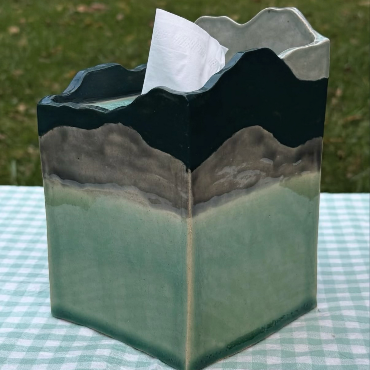 A handmade ceramic tissue box cover shaped like a mountain range, sitting on a green and white checkered tablecloth with a grassy background. The tissue is pulled out of the top opening.