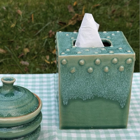 A green glazed ceramic tissue box cover with a tissue insert and a matching lidded container, displayed on a light blue and white checkered tablecloth with grass in the background.