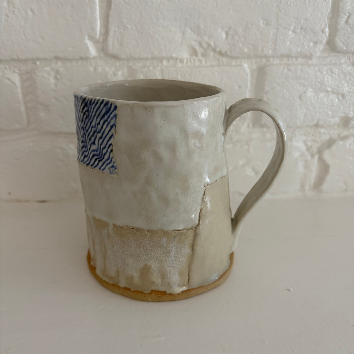 A handmade ceramic mug with a textured, patchwork-style glaze in off-white and beige, featuring a blue patterned accent on the upper left and a brown clay foot ring, set against a white brick wall background.