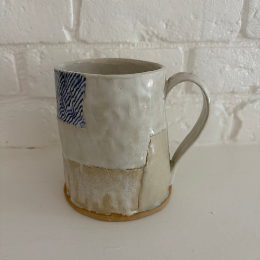 A handmade ceramic mug with a textured, patchwork-style glaze in off-white and beige, featuring a blue patterned accent on the upper left and a brown clay foot ring, set against a white brick wall background.