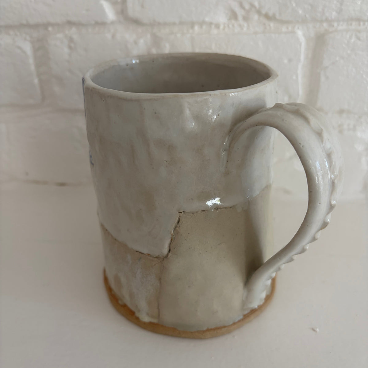 A handmade ceramic mug with a textured, patchwork-style glaze in off-white and light gray, featuring a rustic brown clay base, set against a white brick wall background.